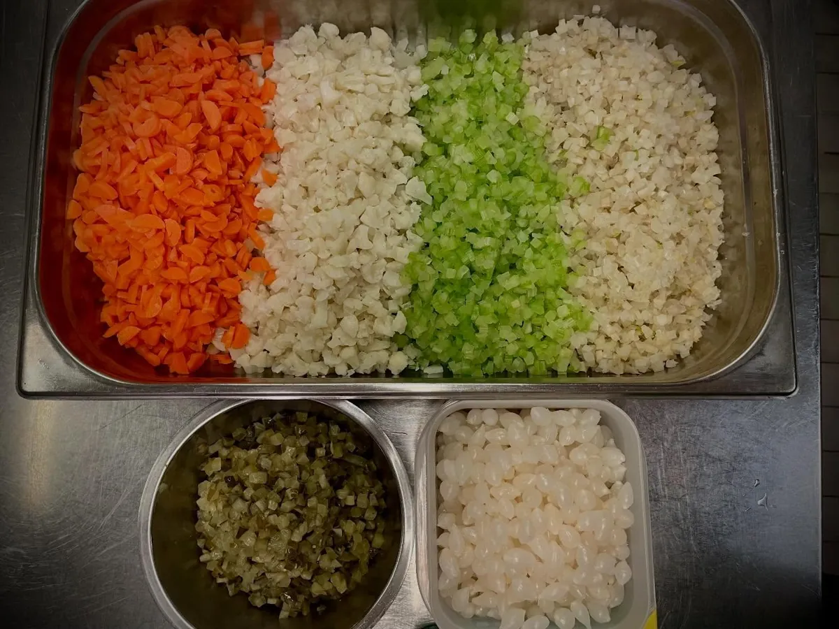 Een bak met fijn gesneden groenten in vier rijen, van oranje wortel tot groene selderij en bloemkool, met daaronder twee kleinere bakjes