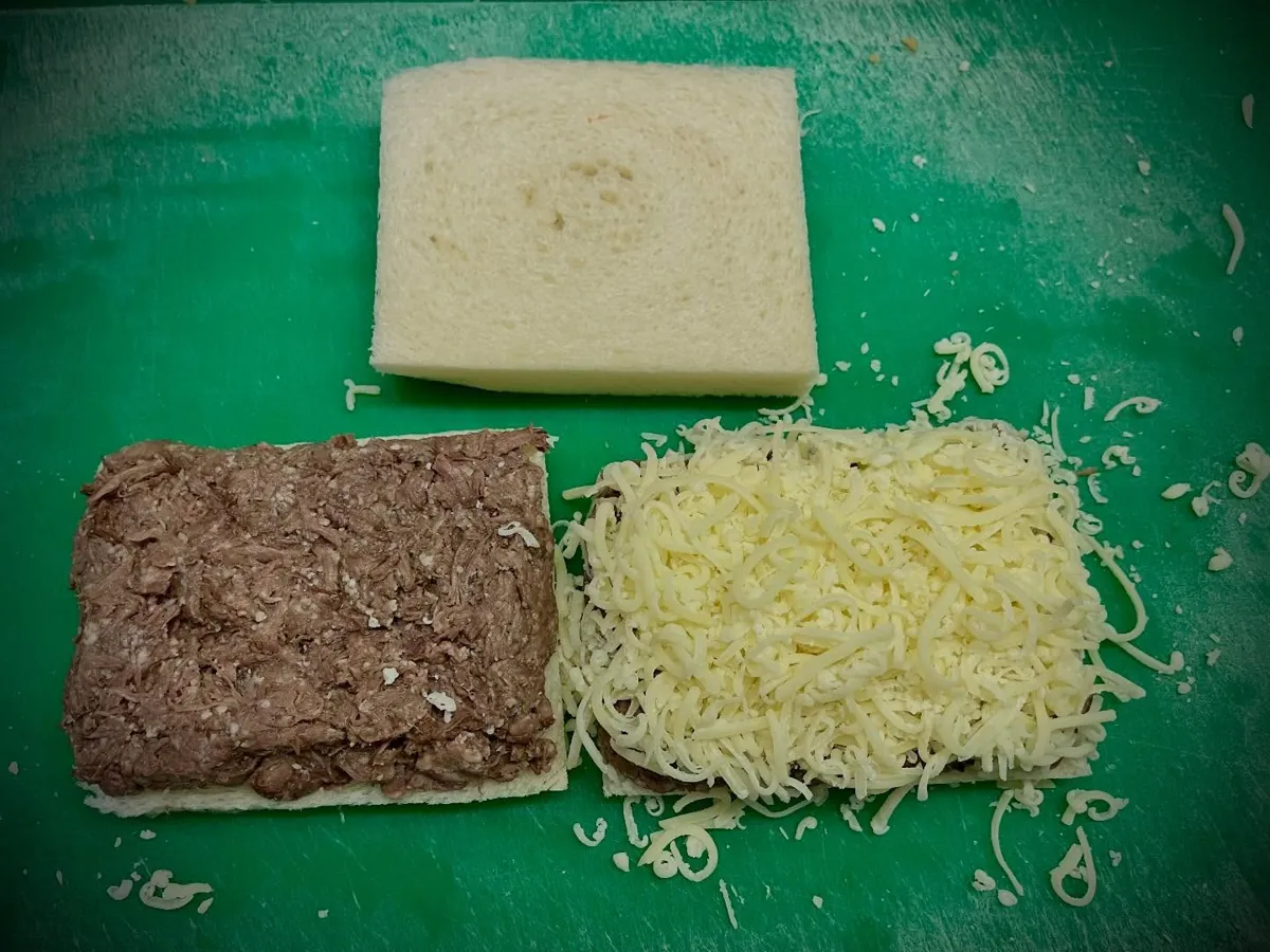 Een snijplank met een boterham met stoofvlees, een hoop geraspte emmentaler en een tweede boterham, klaar om een tosti van te maken